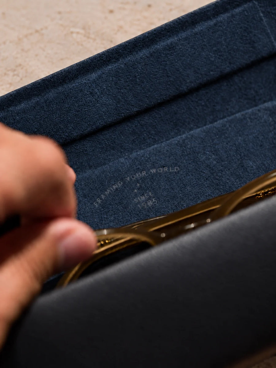 A hand lifting a gold frame out of the open case, showing the navy blue flannelette lining printed with Framing your world, since 1985.